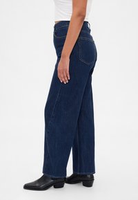 Σκούρα μπλε τζιν παντελόνια με φαρδιά μπατζάκια από τζιν ύφασμα, με ψηλή μέση, πίσω τσέπες και καθαρή ραφή. Συνδυασμένα με μαύρα μποτάκια αστραγάλου.