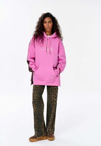 Růžová oversized mikina s přední kapsou, bílým nápisem, spárovaná s hnědými kalhotami ve zvířecím vzoru a hnědými platformovými botami.