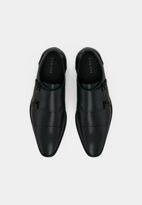 Et par sorte læder monk strap herresko med dobbelte spænder, set ovenfra på en hvid baggrund.
