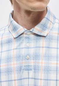 Κοντινό πλάνο ενός άντρα που φοράει ένα λευκό πουκάμισο με κουμπιά και καρό μοτίβο σε μπλε και πορτοκαλί χρώμα με διπλωμένο γιακά.