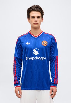 Jeune homme portant un maillot de football bleu à manches longues de Manchester United avec des rayures rouges, le logo Snapdragon et l'emblème blanc d'Adidas, debout sur fond blanc.