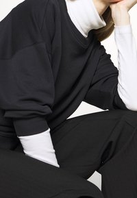 Sudadera negra de gran tamaño con puños acanalados, superpuesta sobre un cuello alto blanco. Presenta un corte amplio, textura suave y un diseño minimalista.