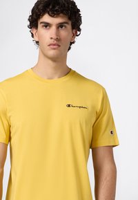 Junger Mann mit lockigem Haar, der ein gelbes Champion-T-Shirt mit Logo auf der Brust und am Ärmel trägt und leicht zur Seite blickt.
