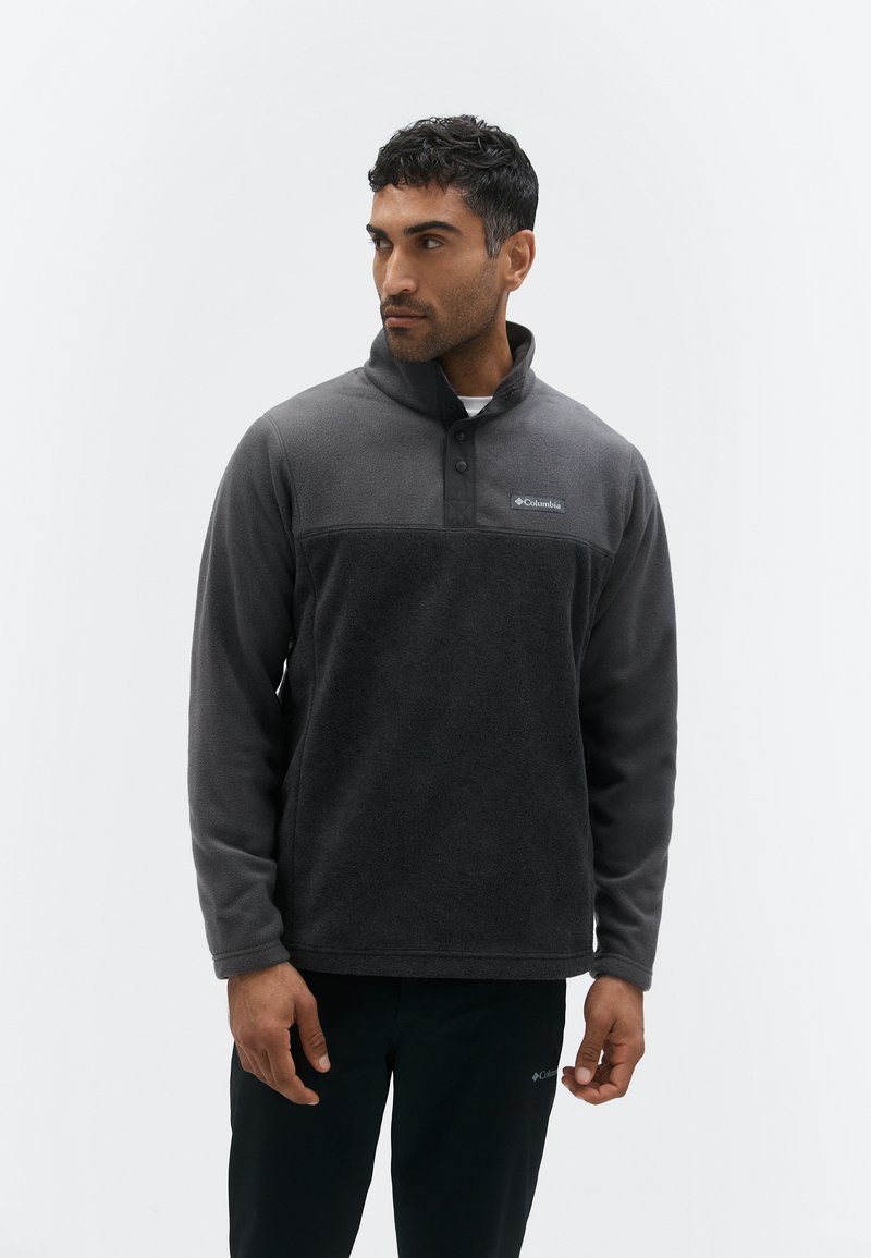 Mann trägt einen dunkelgrauen Fleecepullover mit einem Kragen mit Druckknöpfen und schwarze Hosen, steht vor einem schlichten hellen Hintergrund.