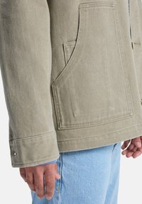 Χακί πράσινο μπουφάν από υφασμάτινη ύφανση, με μεγάλο μεγάλο μπροστινό τσέπη, μανσέτες με κουμπιά και άνετη εφαρμογή, συνδυασμένο με ανοιχτό μπλε τζιν.