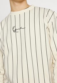 Camisa de manga larga color crema con rayas verticales negras y un pequeño logotipo negro en la zona del pecho. Textura de tejido suave.
