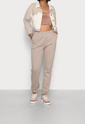 Femme portant une veste courte beige à blocs de couleur, un crop-top mauve, un pantalon de jogging beige et des baskets blanches et roses, debout les mains dans les poches.