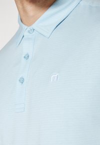 Lyseblå polo skjorte med en teksturert finish, som har en tre-knappers stolpe og en liten brodert logo i hvitt på brystet.