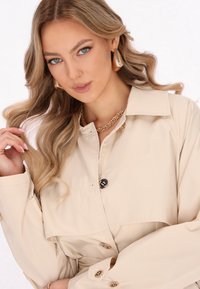Béžový trenčkot s širokým límcem, s dvoubarevným designem a černými knoflíky. Modelka má na sobě zlaté doplňky a vlnité vlasy.