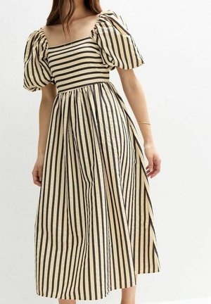 Vrouw draagt een beige jurk met zwarte verticale strepen, met opgeblazen korte mouwen en een vierkante halslijn, staand tegen een effen achtergrond.