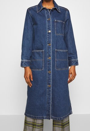 Manteau en denim bleu mi-long avec fermeture à boutons, trois poches plaquées, porté sur un pantalon à carreaux gris avec des rayures jaunes.