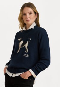 Marineblauwe gebreide trui met een textuur motief van een hond in zwart en wit. Voorzien van een ribbelige ronde hals en manchetten, gedragen over een witte blouse.