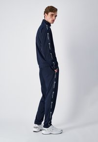 Navy tracksuit set made of smooth fabric, featuring a zip-up jacket and elastic waistband pants. White logo accents down sleeves and pant legs.