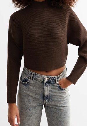 Femme portant un pull court à manches longues côtelé brun foncé et un jean taille haute délavé clair, une main dans la poche.