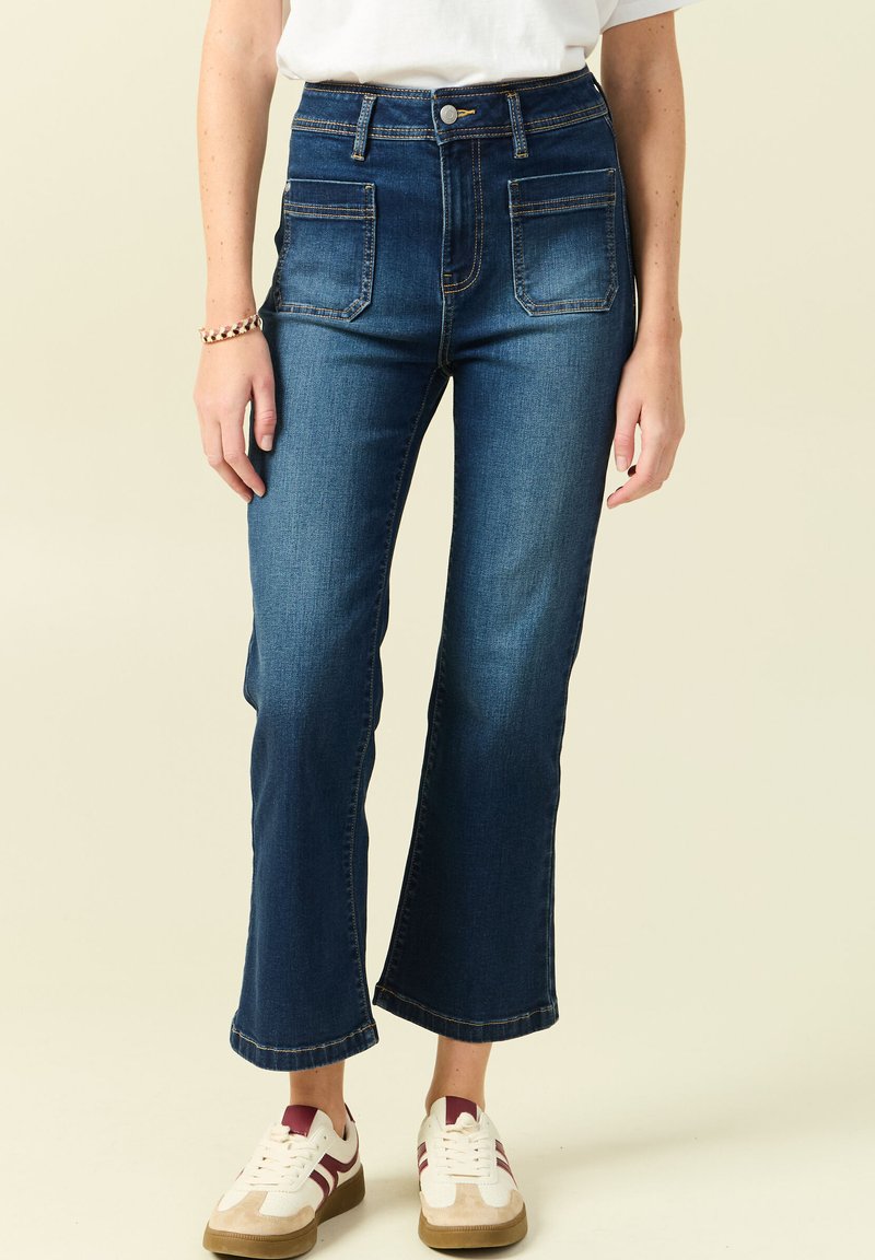 Jeansy z wysokim stanem w ciemnym dżinsie z szerokimi nogawkami, z dwiema przednimi kieszeniami i kontrastowym przeszyciem, zestawione z białymi i bordowymi trampkami.