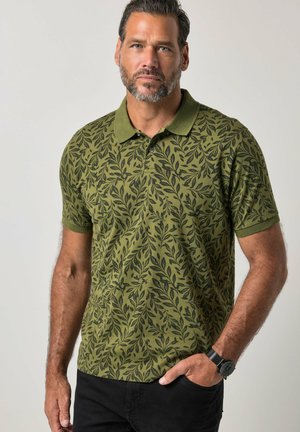 Man met kort donker haar en baard, die een olijfgroene poloshirt met zwarte bladvormen en een zwarte broek draagt, staand met één hand in de broekzak.
