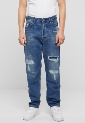 Jean en denim bleu avec une coupe décontractée, ornés d'accents déchirés sur les deux genoux, cinq poches et une fermeture à bouton. Portés avec des baskets blanches.