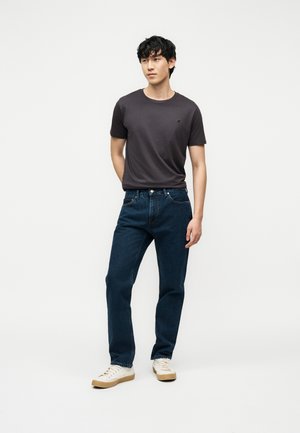 Noormees, kes kannab tumehallist T-särki, tumelisi teksapükse ja valgeid tenniseid, seisab ühe käega selja taga ühevärvilise taustaga.