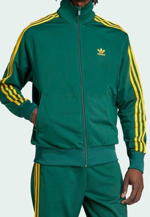 Homme portant un survêtement Adidas vert à rayures jaunes, veste zippée et pantalon assorti, debout avec une main dans la poche.