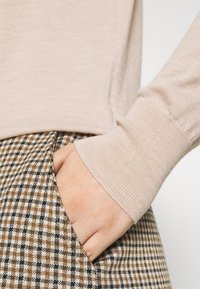 Un pull beige clair avec des poignets côtelés associé à un pantalon à motif écossais marron et noir, montrant une main dans la poche à gauche.