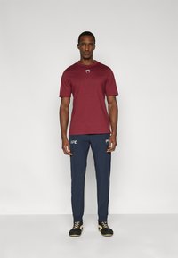 Μπορντό κοντομάνικη κοντομάνικη μπλούζα με λογότυπο, συνδυασμένη με ναυτικοπέδιλα παντελόνια με λευκή σήμανση UFC και μαύρα παπούτσια με κίτρινες κορδόνια.