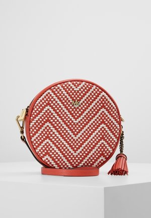 Okrągła torba na ramię z koralowego i białego plecionego materiału z wzorem w jodełkę, złotym logo "MK", bocznym frędzlem i regulowanym paskiem na białej powierzchni.
