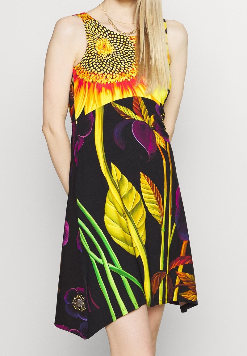 Robe noire sans manches avec un grand imprimé vibrant de tournesol et de feuilles tropicales, portée par une personne aux longs cheveux blonds sur un fond uni.