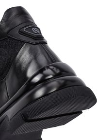 Zapatilla negra con parte superior de cuero suave, panel de tejido texturizado, tacón redondeado y brillante, y suela de goma con un agarre estampado.