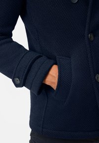 Marinblå texturerad jacka med knäppning framtill, en figursydd krage och en sidficka på vänster ärm med ribbad manschett.