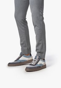 Personne portant un pantalon gris et des baskets décontractées lacées bleues, blanches et brunes, se tenant sur un fond blanc.