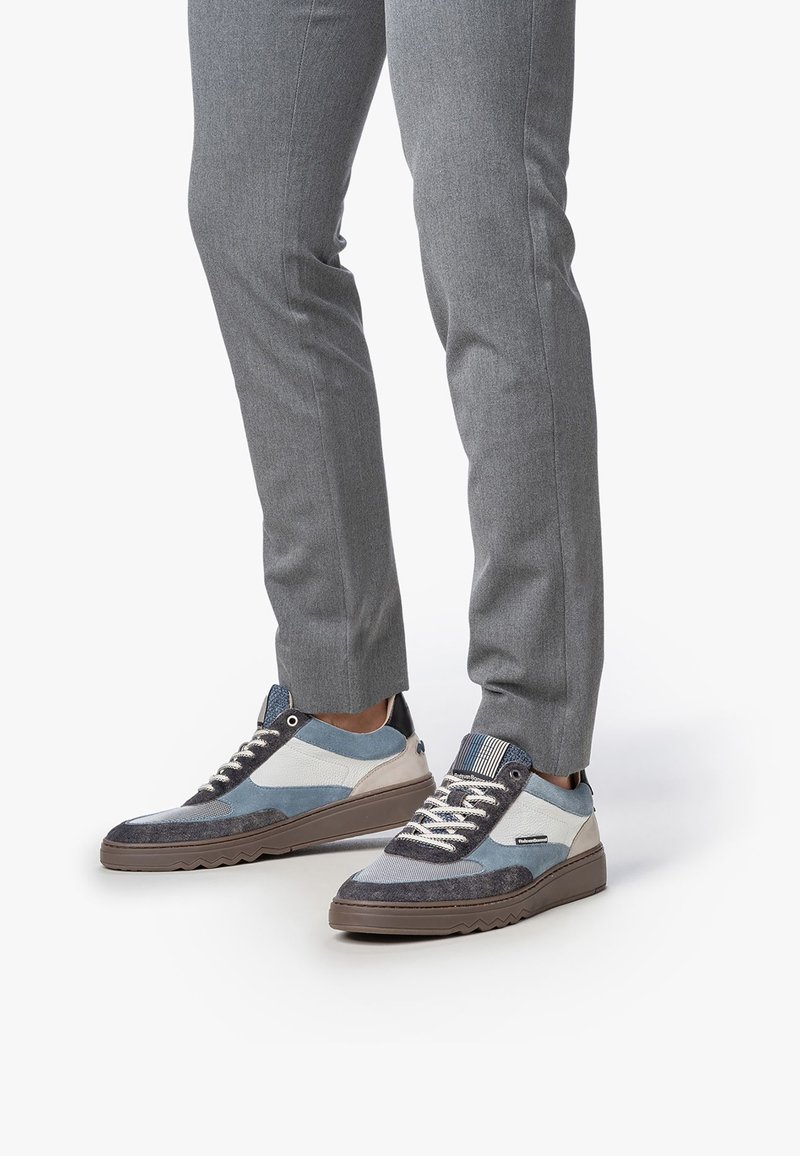 Personne portant un pantalon gris et des baskets décontractées lacées bleues, blanches et brunes, se tenant sur un fond blanc.