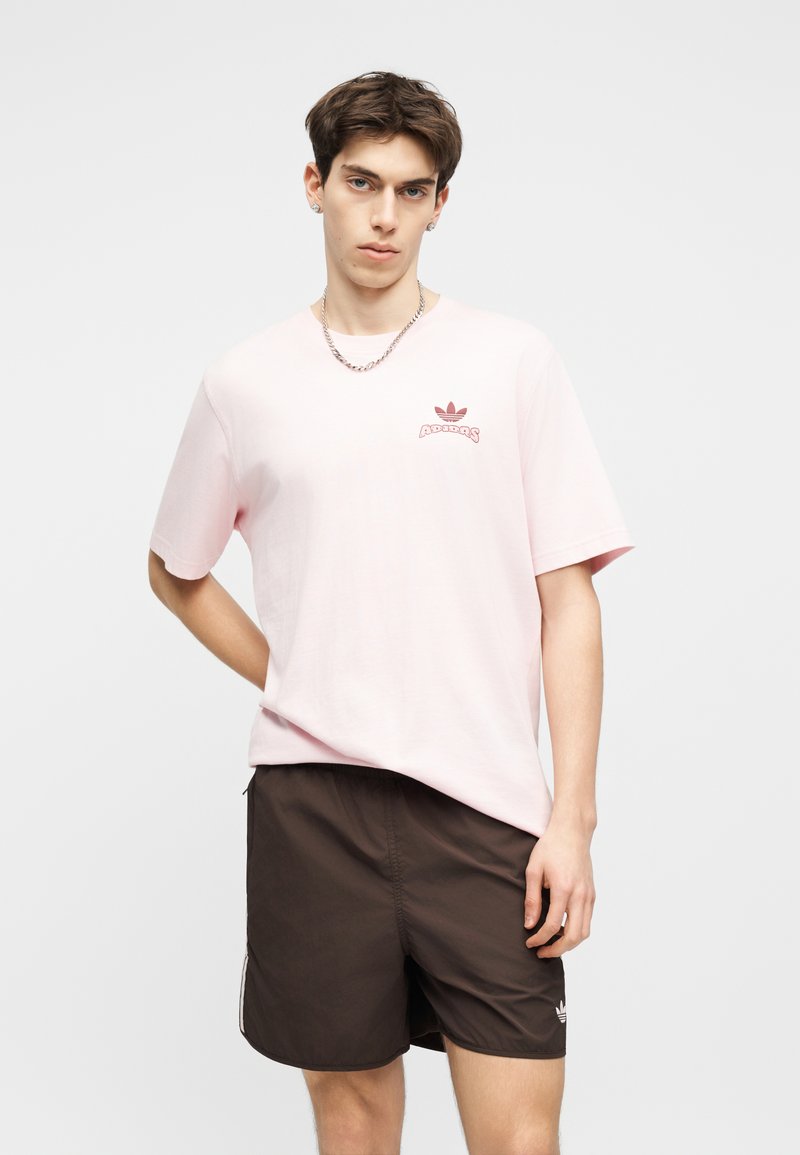 Giovane uomo che indossa una t-shirt Adidas rosa chiaro e pantaloncini Adidas marrone scuro, in piedi davanti a uno sfondo bianco uniforme.