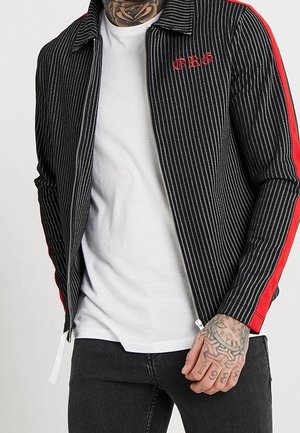 Muž v černém proužkovaném saku s červenými pruhy na rukávech a červeným logem "CRG", v bílé košili, černých džínách a s viditelnými tetováními na rukou.