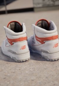 Balti aukšti sportiniai bateliai su oranžinėmis detalėmis ir tekstūruota pilka padais, su "new balance" logotipu ir paminkštintu apvadu.