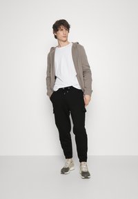 Grijze zip-up hoodie over een wit t-shirt, gecombineerd met zwarte cargobroeken. Schoenen zijn grijze en beige sneakers. Model staat tegen een effen achtergrond.