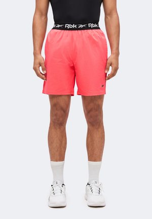 Mann trägt leuchtend pinke Sportshorts mit schwarzem elastischem Bund und weißen Turnschuhen und steht vor einem einfarbigen Hintergrund.