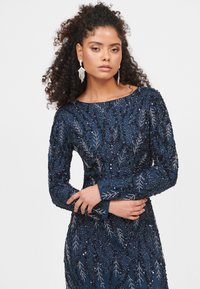 Marineblaues Langarmkleid, verziert mit aufwendiger Perlenarbeit und Blattmustern. Mit tailliertem Schnitt und Rundhalsausschnitt. Strukturierter Stoff mit Schimmer.