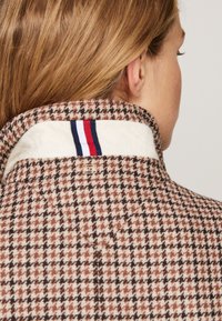 Colar de casaco em padrão houndstooth castanho e branco, com forro creme e uma etiqueta têxtil com riscas vermelhas, brancas e azuis.