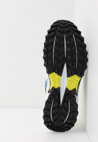 Sportschuhsohle mit tiefschwarzen Traktionsrillen, einem gelben Gummiakzent und einem weißen Obermaterial mit blauen und grünen Details.