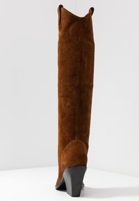 Brauner Wildlederstiefel über dem Knie mit spitz zulaufender Zehe, geschwungener Knöchelnaht und schlankem schwarzem Absatz. Glatte Textur mit matter Oberfläche.