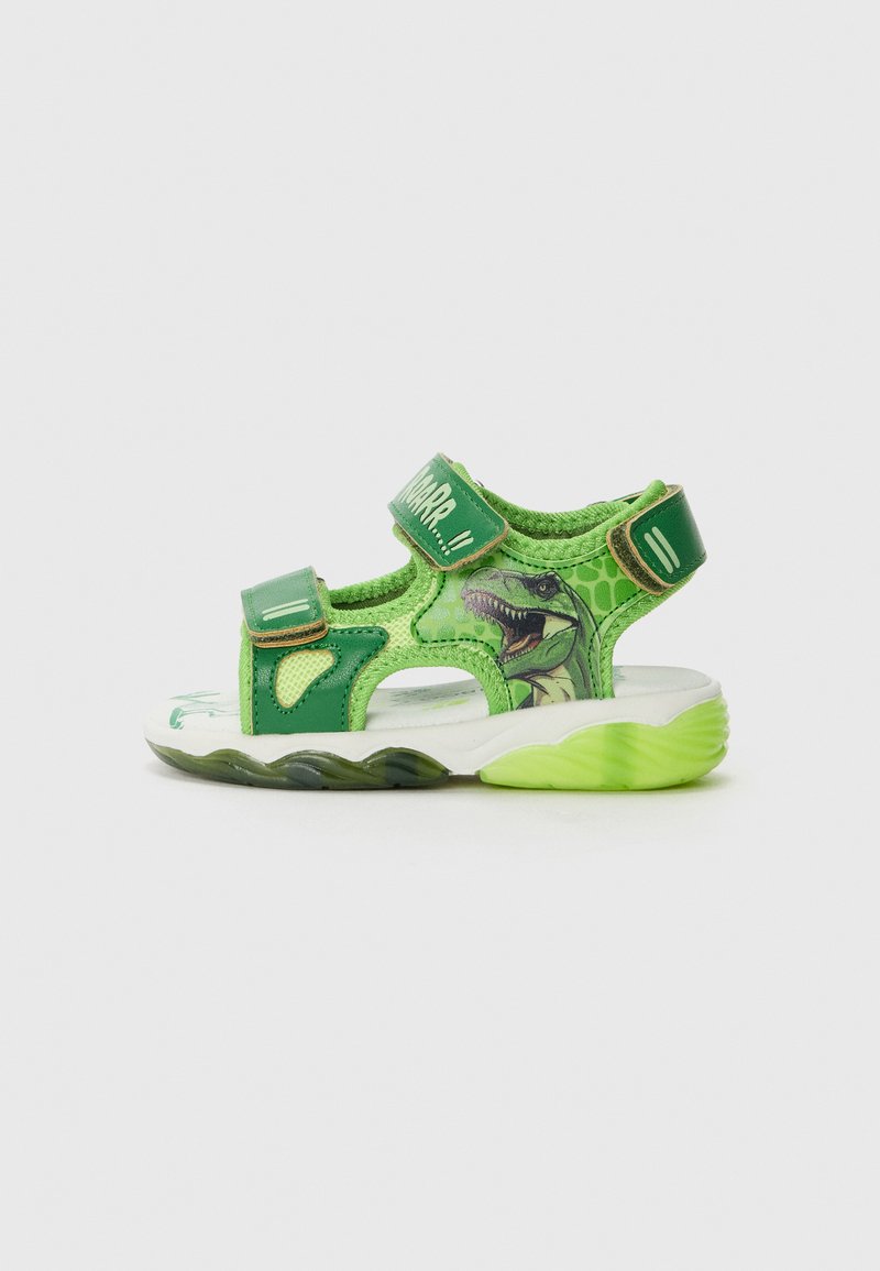 Sandale verte avec des sangles Velcro, ornée d'un motif de dinosaure et d'un dessus texturé. Semelle blanche avec des accents verts et un motif en relief.