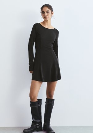 Femme portant une robe noire moulante à manches longues avec une jupe évasée et des bottes hautes en cuir noir, debout contre un mur blanc uni.