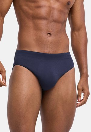 Męski tors ubrany w ciemnoniebieskie slipy, ukazujący zdefiniowane mięśnie brzucha i ud na jednolitym jasnym tle.