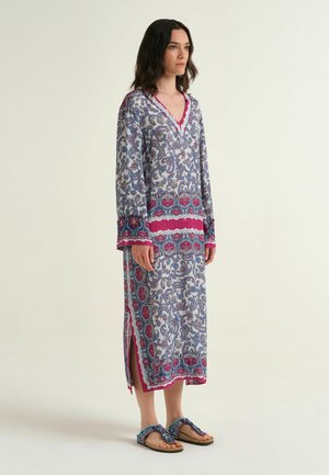 Donna che indossa un lungo vestito con motivo paisley con accenti rosa e blu, in piedi scalza su uno sfondo semplice.