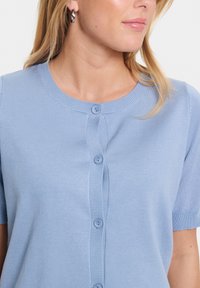 Ljust blå cardigan med rund halsringning, korta ärmar, ribbad textur och knäppning fram. Innehåller två matchande knappar.