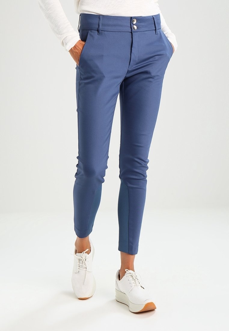 Pantalon bleu marine slim fit avec fermeture à double bouton, associé à des baskets blanches et les mains dans les poches, porté par une personne debout.