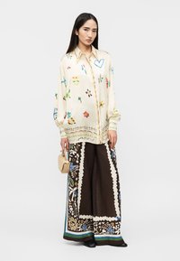 Γυναίκα που φοράει εκρού μπλούζα με πολύχρωμα αφηρημένα σχέδια και φαρδιά παντελόνια με σκοτεινό μοτίβο, κρατώντας μια μικρή μπεζ τσάντα.