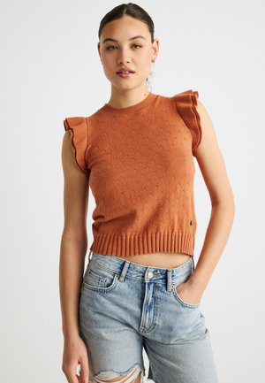 Suéter sin mangas de punto naranja con detalle de hombros con volantes, presenta un patrón texturizado y dobladillo acanalado, combinado con pantalones cortos de mezclilla desgastados.