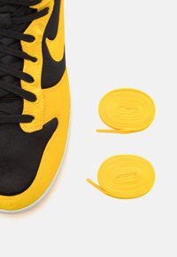 Närbild av en gul och svart sneakersko med svarta skosnören, tillsammans med två prydligt spiralvridna gula skosnören på en vit bakgrund.