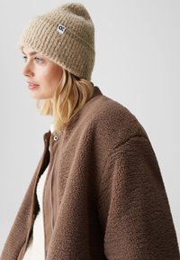 Beige ribbed knit beanie with a logo patch, paired with a brown fluffy jacket featuring a zip closure and textured fabric.